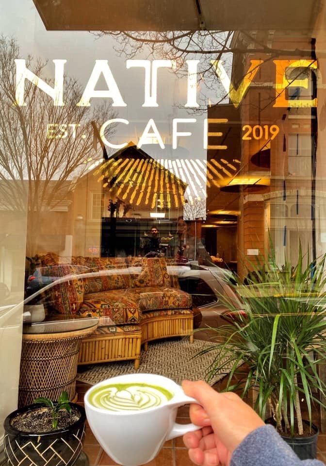 Warm sunlight-filled Native Cafe interior with wooden tables, lush green plants, and a professional espresso machine